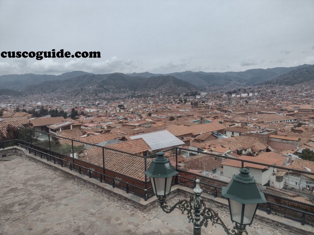 Panoramic view of downtown Cusco from the San Blas viewpoint, showing the historic city center, red rooftops, colonial buildings, and surrounding Andean mountains.
